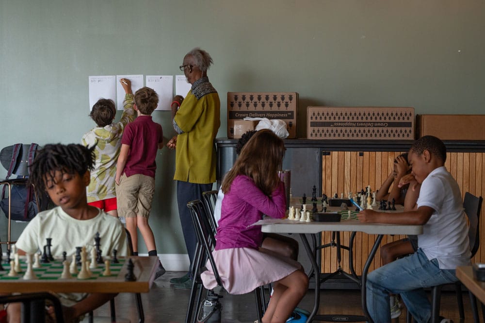 Groups of children sit at tables playing games of chess. Two children are seen noting their scores on sheets posted to the wall.