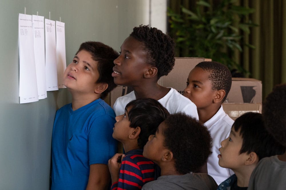 A group of children looks at chess moves on sheets posted to the wall.