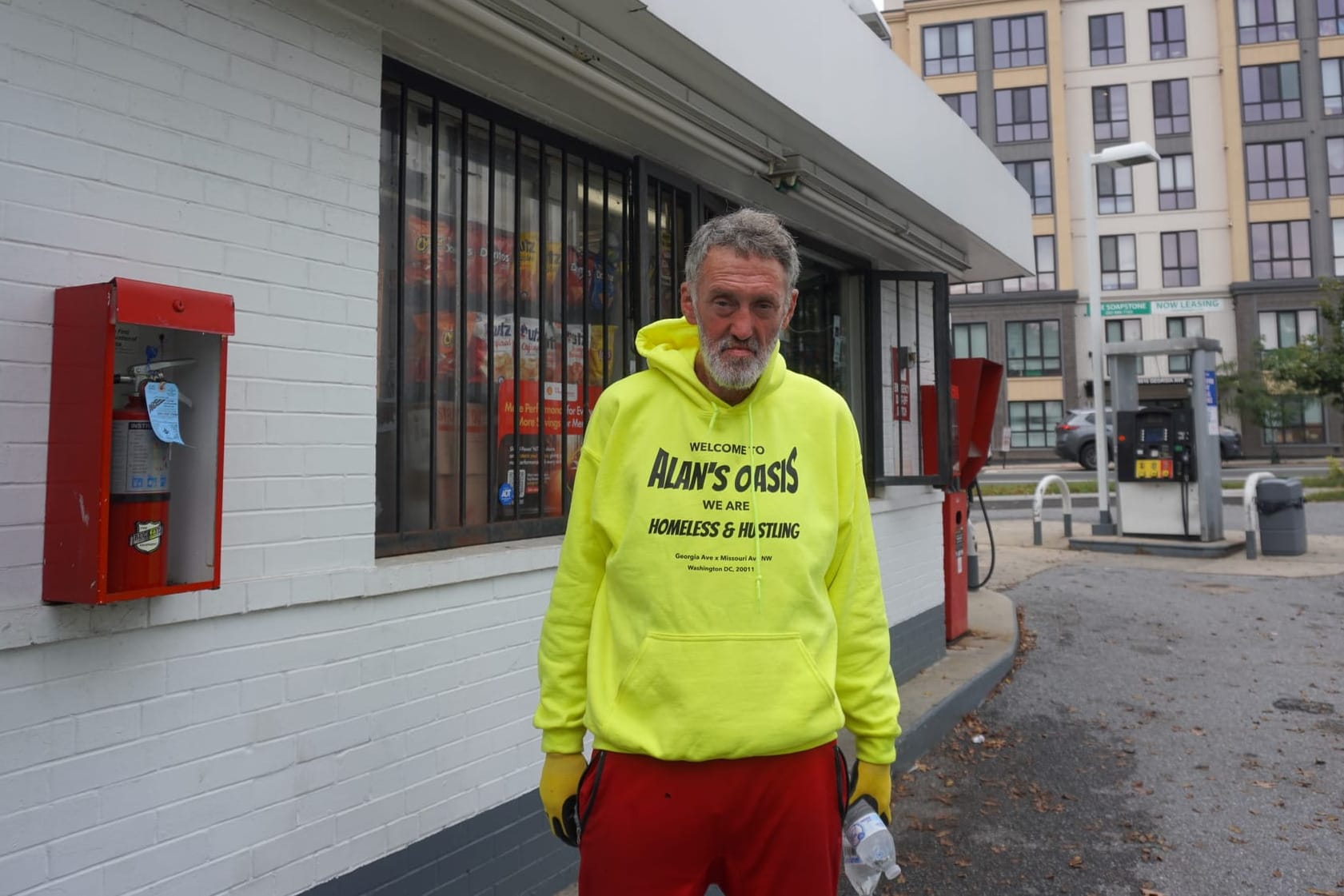 A man at a gas station wears a highlighter yellow sweatshirt reading "Welcome to ALAN's OASIS / We are homeless & hustling"