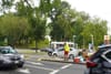 A person stands on the median of a busy street, next to a board propped up against coolers and a shopping cart, as cars pass by